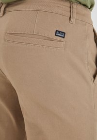 Pantalons beiges en tissu mélangé de coton, dotés d'une poche arrière avec un accent cousu et d'une petite étiquette de marque. Texture lisse.