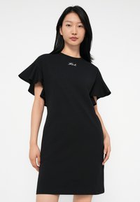 Νέα γυναίκα φορώντας ένα μαύρο φόρεμα μέχρι το γόνατο με φουσκωτά μανίκια και ένα μικρό λευκό λογότυπο σε στυλ γραφής στο στήθος, που στέκεται μπροστά από ένα απλό φόντο.