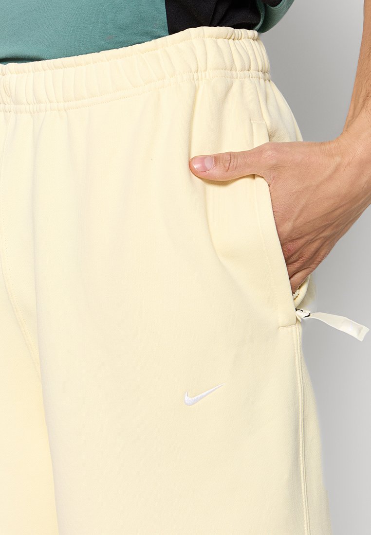 Pantalones de chándal de color amarillo claro con cintura elástica, bolsillos laterales y un pequeño logo blanco de Nike en la pierna izquierda. Textura de tela suave.