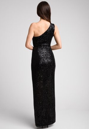Kobieta z długimi brązowymi włosami, ubrana w czarną, cekinową suknię o długości do ziemi z jednym ramiączkiem, stojąca plecami w kierunku patrzącego, z rękami na biodrach.