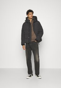 Jachetă puffer neagră cu design matlasat, glugă cu șnur, purtată peste un top maro, asociată cu blugi negri destrămați și adidași negri.