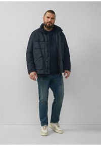 Marineblauwe jas met meerdere zakken, knopenaccenten en een capuchon, gecombineerd met een donker shirt en versleten blauwe jeans, en lichtgekleurde sneakers.