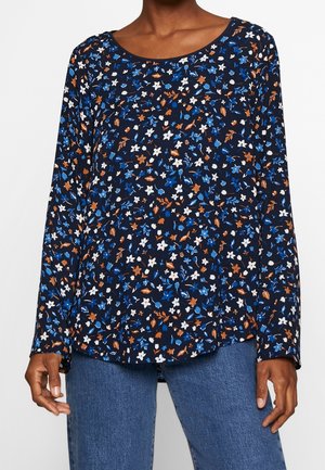 Frau trägt eine langärmelige marineblaue Bluse mit kleinen blauen, weißen und braunen Blumenmustern, kombiniert mit blauen Jeans aus Denim.