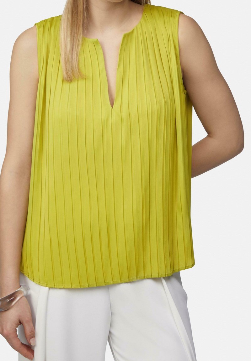 Femme portant un chemisier plissé jaune sans manches à encolure fendue et un pantalon blanc, sur fond uni.
