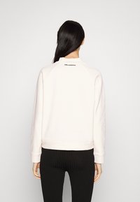 Žena s dlouhými černými vlasy má na sobě světle růžovou mikinu s nápisem "KARL LAGERFELD" poblíž krku a černé žebrované kalhoty, stojí zády.