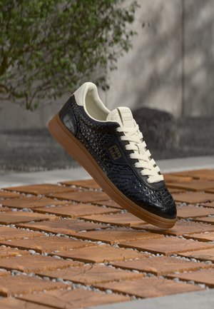Baskets noires texturées avec lacets blancs et semelle blanche, flottant au-dessus d’un sol en carreaux de bois avec une plante verte floue en arrière-plan.