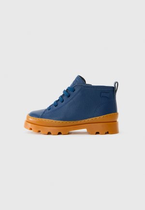 Botte en cuir bleue à la cheville avec des lacets bleus et une semelle épaisse à motif orange, vue de côté sur un fond gris clair.