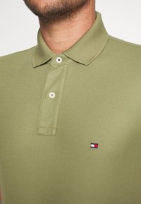 Gros plan sur un homme portant un polo vert clair avec une patte de boutonnage à deux boutons et un petit logo rouge, blanc et bleu sur la poitrine.