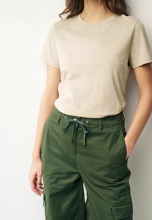 Kvinde iført beige kortærmet skjorte stoppet ned i højtaljede grønne cargobukser med snøre og hånd i lommen.