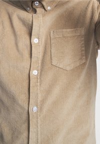 Chemise en velours côtelé tan avec une texture en côtes verticales, boutons blancs et une poche sur la poitrine. Le tissu semble doux et légèrement texturé.