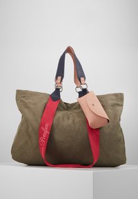 Große olivgrüne Canvas-Tasche mit navyfarbenen Ledergriffen und rotem Riemen, mit einer kleinen rosa Lederpouch, die angehängt ist. Text auf dem Riemen gestickt.