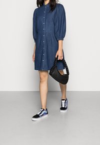 Denim shirt dress with puff sleeves and front buttons; paired with black braided handbag and black sneakers featuring blue accents.