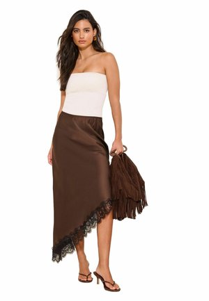 Vrouw draagt een strapless witte top, asymmetrische bruine rok met kanten rand, bruine handtas met franjes en bruine sandalen met hakken, staand tegen een witte achtergrond.