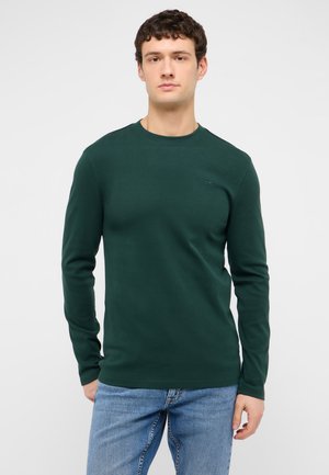 Man wearing a dark green long-sleeve shirt and blue jeans standing against a plain white background.