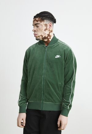 Veste zippée en velours vert avec des bandes latérales blanches et un petit logo blanc Nike sur la poitrine. Dotée d'un col montant et de poches latérales.