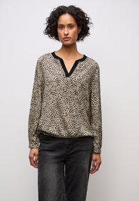 Kobieta o kręconych włosach, ubrana w beżową, czarno nakrapianą bluzkę z długim rękawem, z dekoltem w kształcie litery V wykończonym czarną lamówką oraz wyblakłe czarne jeansy na jednolitym tle.