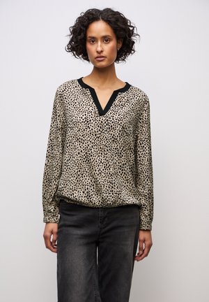 Frau mit lockigem Haar, die eine beigefarbene, schwarz gefleckte Langarmbluse mit schwarz abgesetztem V-Ausschnitt und ausgewaschene schwarze Jeans vor einem einfarbigen Hintergrund trägt.