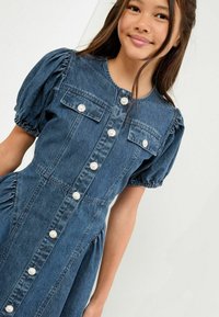 Rifľové šaty s krátkymi nafúknutými rukávmi, dvoma náprsnými vreckami a gombíkmi pripomínajúcimi perly. Tkanina má klasickú modrú rifľovú textúru.