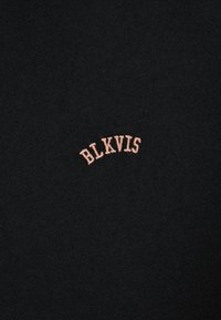 Textil negro con un logotipo bordado en forma curva que dice "BLKVIS" en hilo rosa claro, vista en primer plano que enfatiza la textura y los detalles.