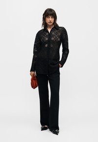 Γυναίκα που φοράει μαύρη φλοράλ δαντελένια μπλούζα με μακριά μανίκια, μαύρο παντελόνι με φαρδιά μπατζάκια, μαύρα μυτερά παπούτσια, κρατώντας μια μικρή καφέ τσάντα φάκελο.