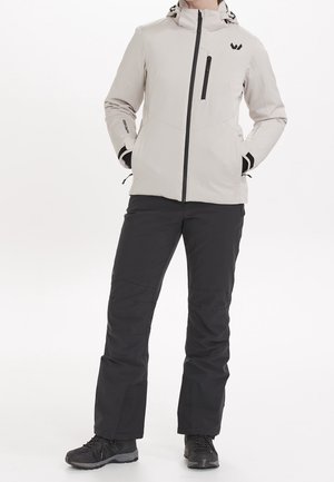 Persoon draagt een lichtgrijze capuchonjas met zwarte rits en zakken, zwarte broek en zwarte wandelschoenen, staand met handen in de zakken.