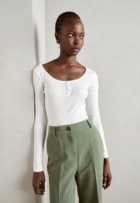 White long-sleeve top with a scoop neckline and three-button placket, paired with high-waisted olive green trousers featuring a button closure.