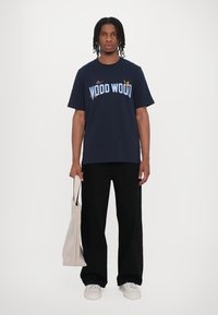 Camiseta de algodón azul marino con gráfico "WOOD WOOD" en azul claro, combinada con pantalones de pierna ancha negros y una bolsa de lona beige.