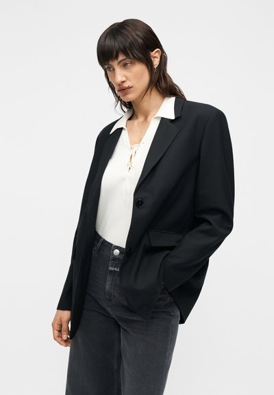 Μαύρο σακάκι με μία κουμπί, μεγάλο πέτο με σχισμή, φορεμένο πάνω από λευκό ριμπ μπλουζάκι και γκρι τζιν ψηλόμεσα παντελόνια.