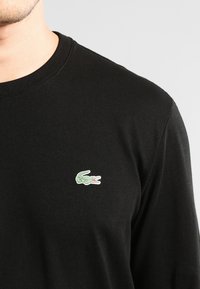 Black long-sleeve t-shirt made of cotton with a round neckline. Features a small green embroidered crocodile logo on the chest.
