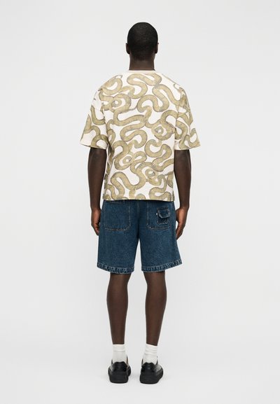 Πουκάμισο με κοντά μανίκια σε μπεζ χρώμα με σχέδια σε κίτρινο και πράσινο που δημιουργούν στροφές, συνδυασμένο με μπλε τζιν σορτς και μαύρα παπούτσια.