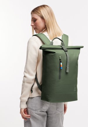 Donna bionda che indossa un maglione crema e jeans grigi, porta uno zaino verde con chiusura a rotolo e tasca frontale con cerniera.