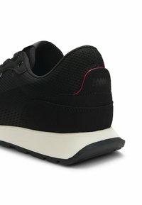 Baskets athlétiques noires avec dessus en maille, accent rouge sur la languette arrière, semelle intermédiaire blanche et semelle extérieure noire visible depuis un angle arrière latéral.