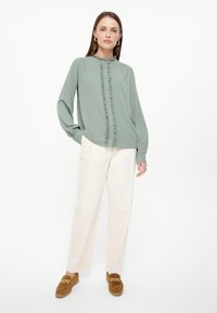 Blusa de manga larga verde claro con bordes con volantes y botones en el frente, combinada con pantalones de pierna ancha color crema y zapatos tipo slip-on marrones.