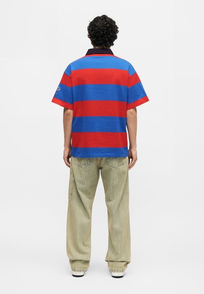 Άνδρας με σγουρά μαλλιά, φοράει ένα κόκκινο και μπλε πουά πόλο πουκάμισο και φαρδιά ανοιχτό μπεζ παντελόνια, στέκεται με την πλάτη προς τα εμπρός σε απλό φόντο.