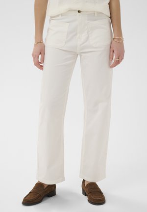 Žena oblečená v bielych nohaviciach rovného strihu s prednými nášivkami a hnedými semišovými mokasínami, zobrazená od stredu trupu po nohy.