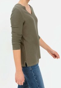 Oliwkowa dzianinowa bluzka z długim rękawem, z dekoltem w kształcie litery V, bocznymi rozporkami i teksturowanym wykończeniem, zestawiona z niebieskimi dżinsami.