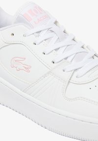 Fehér bőr sportcipő, kerek orral, texturált részletekkel és rózsaszín Lacoste logóval az oldalon. Fehér fűzőkkel és párnázott nyelvvel rendelkezik.