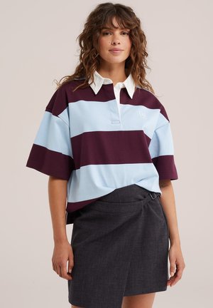 Femme portant un maillot de rugby ample avec de larges rayures bleu clair et violet foncé et une jupe portefeuille gris foncé.