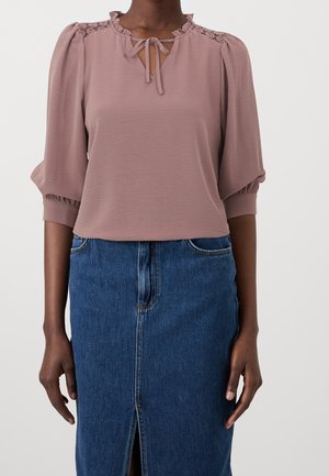 Dunkelhäutige Person trägt eine mauve Bluse mit Spitzen-Details und einer Schleife im Ausschnitt, kombiniert mit einem hoch taillierten blauen Jeansrock mit Frontschlitz.