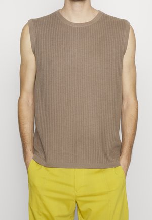 Bez rękawów jasnobrązowy prążkowany dzianinowy top noszony z jasnożółtymi spodniami, dłonie w kieszeniach, tułów widoczny od ramion do bioder.