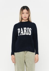 Námornícky sveter s bielym nápisom "PARIS" na prednej strane, okrúhly výstrih, dlhé rukávy, vyrobený z jemnej tkaniny. Skombinovaný s svetlými nohavicami.