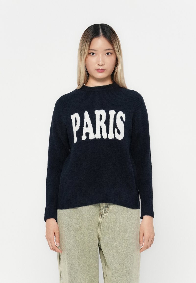 Námornícky sveter s bielym nápisom "PARIS" na prednej strane, okrúhly výstrih, dlhé rukávy, vyrobený z jemnej tkaniny. Skombinovaný s svetlými nohavicami.