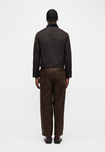 Άνδρας με την πλάτη στραμμένη φοράει ένα σκούρο καφέ μπουφάν, καφέ παντελόνια και σκούρα παπούτσια, μπροστά σε απλό λευκό φόντο.