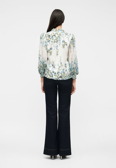 Γυναίκα με μακριά σκούρα μαλλιά που φορά λουλουδάτο μπλουζάκι με μακριά μανίκια και σκούρα, φαρδιά παντελόνια, στέκεται γυρισμένη με την πλάτη προς τα εμπρός.