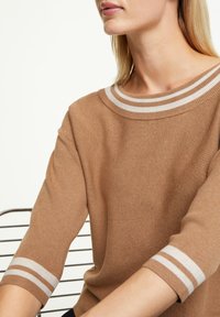 Pull en maille marron avec un large col rayé et des manches trois-quarts ornées de rayures blanches aux poignets. Texture lisse, design décontracté.