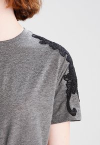Camiseta gris de manga corta con detalles de encaje floral negro en los hombros. Tejido suave con un ajuste regular, que presenta un escote redondo.