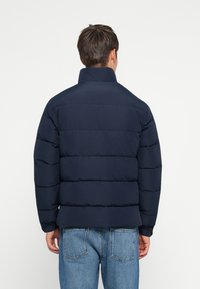 Man wearing navy blue padded jacket and light blue jeans, standing facing away against plain white background.