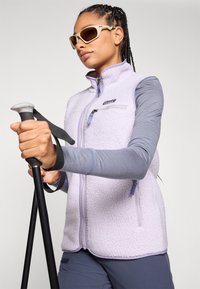 Violet fleeces vestes ar teksturētu virsmu, ar rāvējslēdzēja kabatām un atstarotājiem. Modelis tur pārgājienu nūjas, valkājot saulesbrilles ar bēšiem rāmiem.
