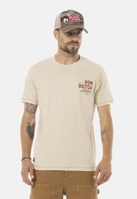 Camiseta beige de manga corta con un pequeño logo marrón de "Von Dutch" en el lado izquierdo del pecho. Se lleva con una gorra de malla beige que muestra gráficos coloridos.