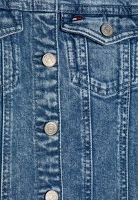 Denimjakke i lys blå med sølvfargede trykknapper, vertikale sømmer og en liten brystlomme. Merker synlige inni jakken.
